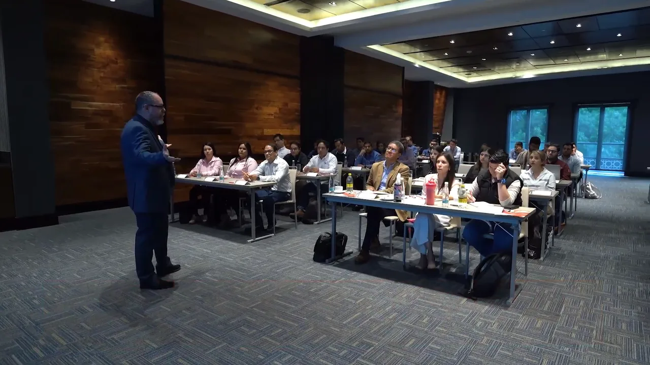 Enrique Vidal de pie delante de una sala de formación con los participantes sentados en mesas, fotograma del vídeo del curso de visitas